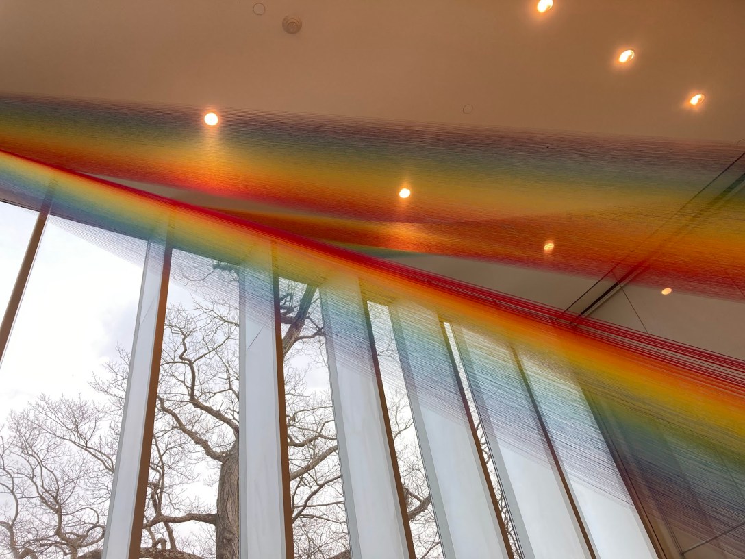 Strings in rainbow colors spread across the Bruce Museum ceiling from the edge of a window, almost like a real rainbow going through a prism.